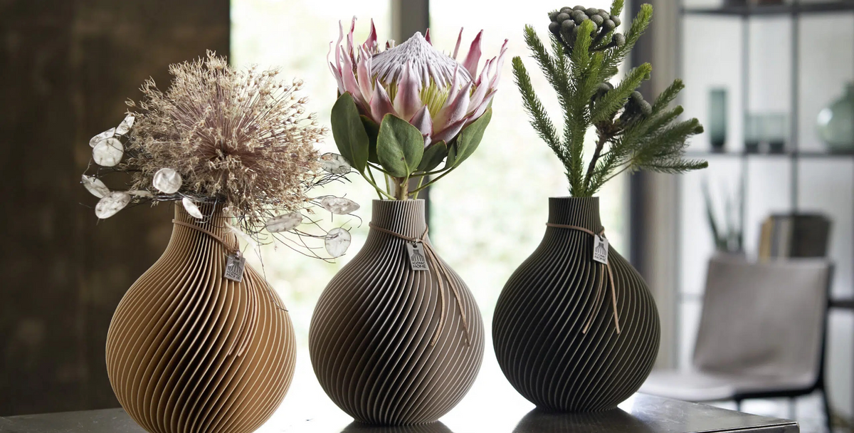 Tre vasi Sphere in natural oak, cozy greige e mystic mud, ognuno con una diversa composizione floreale.